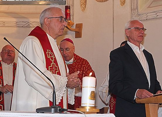Ein Priester leitet eine Zeremonie mit einer brennenden Kerze vorne, während ein anderer Priester neben ihm steht und ein Mann im Anzug zusieht.