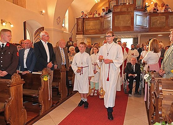 In einer Kirche gehen zwei Jungen in weißen Roben den Gang hinunter. Viele Menschen stehen und sitzen auf Kirchenbänken, und zwei Männer in Anzügen beobachten vom Balkon aus.