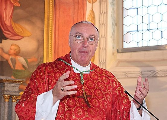 Ein Priester steht vor einem Mikrofon und spricht, er trägt einen roten und weißen Umhang mit einer grünen Schnur um den Hals.
