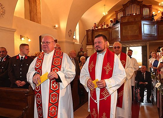 Zwei Priester stehen in einer Kirche, dem Publikum zugewandt, mit einer Galerie im Hintergrund. Der Priester auf der linken Seite hält ein gelbes Buch, während der auf der rechten Seite ein gelbes Objekt hält. Hinter ihnen sitzen und stehen Menschen auf Bänken.