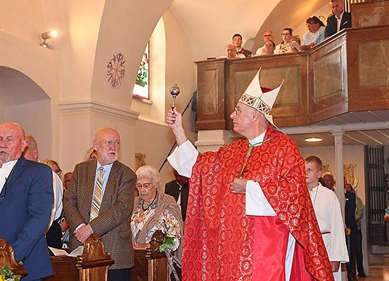 Ein Priester in einer roten Robe hält eine Glocke und leitet eine Zeremonie in einer Kirche. Vor ihm stehen mehrere Menschen, und andere befinden sich auf der Empore.