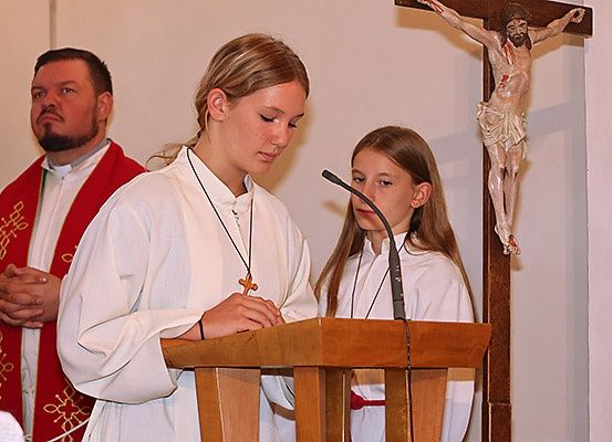 Zwei Mädchen in weißen Gewändern lesen von einer Kanzel in einer Kirche, ein Priester in einem roten Gewand steht hinter ihnen.