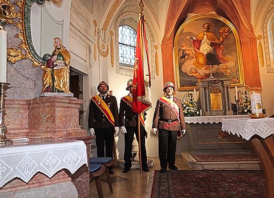 Vier Feuerwehrleute in Uniform stehen in einer Kapelle, halten eine Fahne und stehen vor einem Altar. Hinter ihnen zieren eine Statue und ein Gemälde die Wände.
