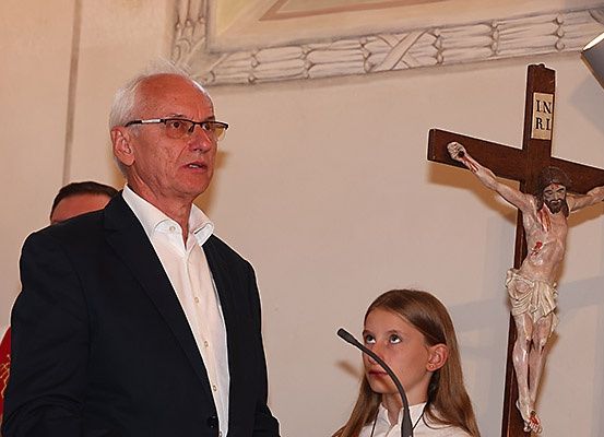 Ein älterer Mann mit Brille steht an einem Rednerpult mit Mikrofon und spricht zu einer Menschenmenge. Ein Mädchen liest neben ihm aus einem Buch. Hinter ihnen ist ein Kruzifix an der Wand angebracht.