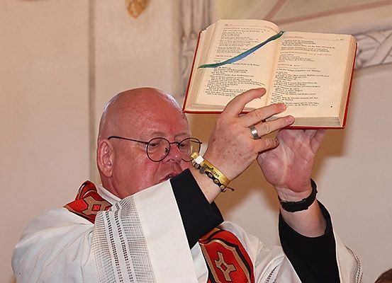 Ein Priester in einer weißen Robe hält ein Buch, möglicherweise ein Gesangbuch, in seinen erhobenen Händen. Er trägt eine Brille, einen Ring und ein Armband.