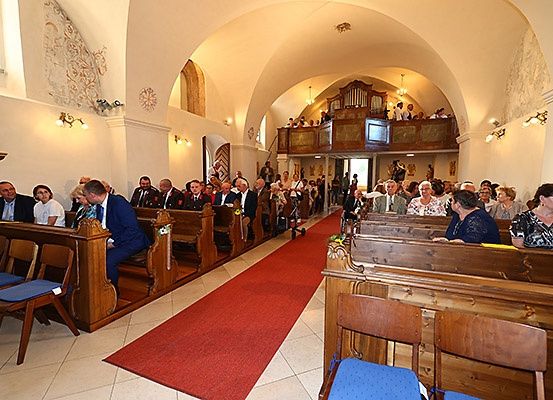 Eine Kircheninnenraum mit hölzernen Kirchenbänken, einem roten Teppich und einer Statue an der Wand. Menschen sitzen auf den Kirchenbänken, und einige stehen in der Nähe des Altars.