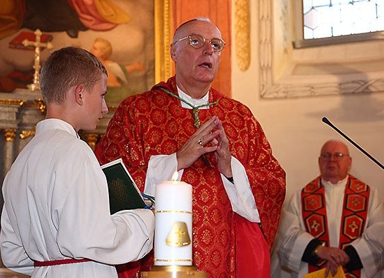 Ein Priester ist dabei zu sprechen in einer Kirche, ein junger Junge hält in der Nähe eine Kerze.