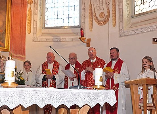 Fünf Priester in roten Gewändern stehen am Altar einer Kirche, halten Kerzen und singen. Eine Frau in einem weißen Gewand steht in der Nähe.