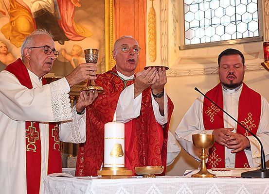 Drei Priester in roten Gewändern halten Kelche, einer mit einer brennenden Kerze, in einer Kirche. Eine Kerze und ein Buch liegen auf dem Altar.