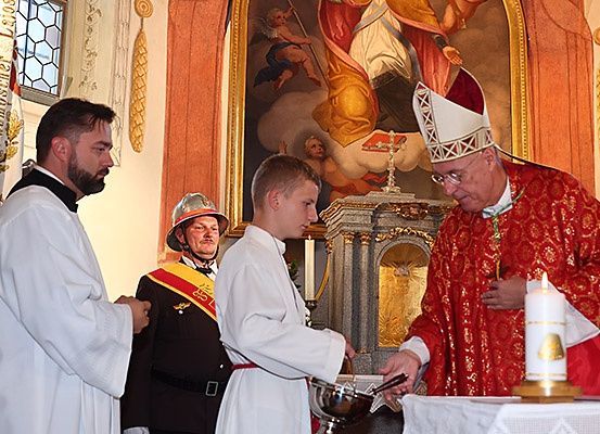 Ein Bischof in einer roten und weißen Robe hält eine Kerze, während ein Mann in einer weißen Robe eine silberne Schale hält. Hinter ihnen steht ein Mann in einer schwarzen Uniform und einem Helm in der Nähe einer Wand mit einem großen Gemälde.