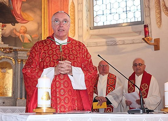 Ein Priester steht in einer Kirche an einer Kanzel, trägt einen roten Roben und eine Brille. Zwei weitere Priester stehen hinter ihm, einer hält ein Mikrofon und der andere ein Stück Papier. Kerzen sind auf dem Tisch angezündet.