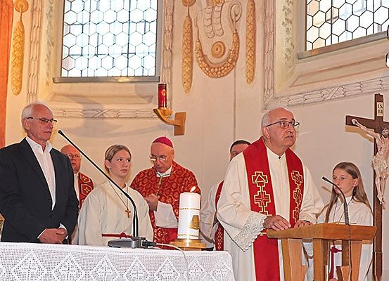 In einer Kirche hält ein Priester am Altar eine Rede, eine Kerze ist angezündet. Hinter ihm stehen zwei Frauen, ein Mann und ein Bischof.