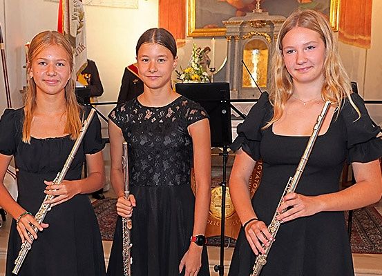 Drei junge Frauen stehen in einer Kirche und halten Flöten. Sie sind in Schwarz gekleidet. Hinter ihnen steht ein Notenständer mit einem Mikrofon. Im Hintergrund sind eine Flagge und eine Statue zu sehen.