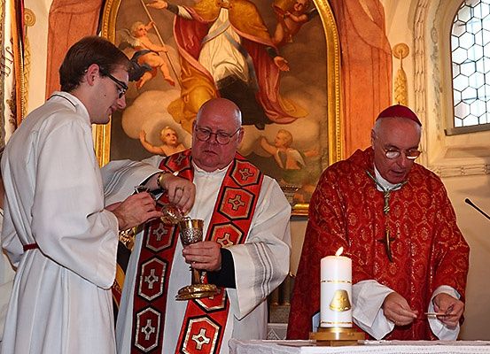 Priester in liturgischen Gewändern führen eine Zeremonie an einem Altar durch, einer hält einen Kelch und der andere zündet eine Kerze an. Ein großes Gemälde im Hintergrund zeigt Engel und eine heilige Figur.
