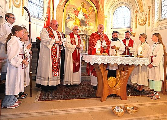 Eine Gruppe von Menschen in weißen Gewändern steht vor einem Kirchenaltar mit Kerzen und einem goldenen Kelch. Priester in roten Gewändern beten und halten ein Mikrofon.