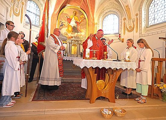 Ein Priester in roten Gewändern führt eine Zeremonie in einer Kirche mit zwei Akolythen und mehreren Kindern durch. Der Altar hat eine Goldmünze, Kerzen und ein Kreuz. Dahinter hält eine Person eine Fahne.