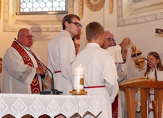 Eine religiöse Zeremonie findet in einer Kirche statt. Ein Mann mit einer Glocke steht an einer Kanzel, vor ihm ein anderer Mann. Ein weiterer Mann hält eine Kerze auf dem Altar.