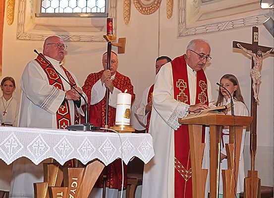Ein Priester in einer rot-weißen Robe liest an einem hölzernen Pult in einer Kirche vor, mit Kerzen und einem Kreuz in der Nähe.
