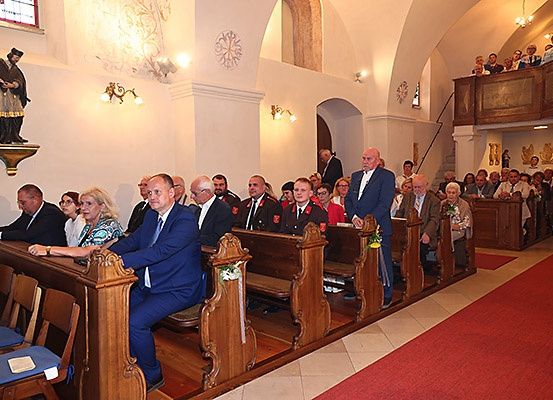 Men und Frauen in formeller Kleidung sitzen auf Kirchenbänken in einer Kirche, mit einer Statue an der Wand und einem roten Teppich, der zum Altar führt.