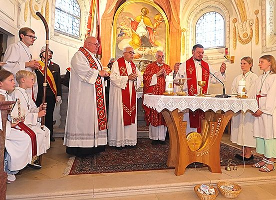 Priester in zeremonieller Kleidung stehen vor einem Altar in einer Kirche, wobei Kinder und Erwachsene beobachten, und ein religiöses Gemälde im Hintergrund.
