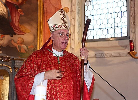 Ein Bischof in roter Robe und mit Brille spricht in einer Kirche, eine Kerze und einen Stab haltend. Hinter ihm befindet sich ein Gemälde und ein Fenster.