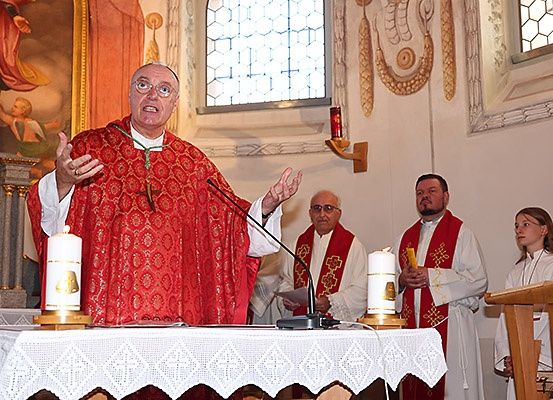 Ein Priester in roter Robe und mit Brille spricht in ein Mikrofon an einem Kirchenaltar.