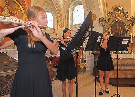 Drei Frauen spielen Flöten in einer Kirche. Sie tragen schwarze Kleider und haben Notenständer vor sich.