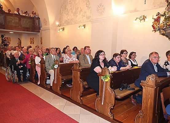 Eine Gruppe von Menschen in einer Kirche, einige in Rollstühlen, die auf hölzernen Kirchenbänken sitzen, mit Blumen geschmückt, und zwei Personen in formeller Kleidung am Gang.