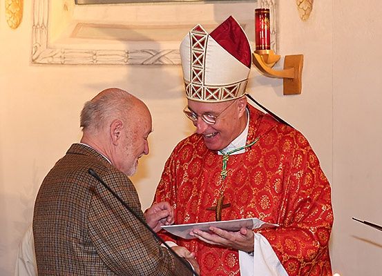 Ein Bischof in einem roten und weißen Gewand übergibt einem Mann in einem braunen Mantel ein Dokument, der vor einem Mikrofon in einer Kirche steht.