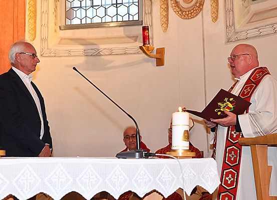 Zwei Männer in einer Kirche, einer mit Brille liest aus einem Buch, der andere in einem Anzug hört zu. Kerzen und ein Mikrofon auf einem Tisch.
