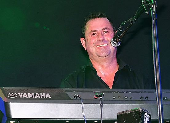 Ein Mann steht hinter einem Yamaha-Keyboard, lächelt und spielt ein Musikinstrument in einem schwach beleuchteten Bereich. Er trägt ein schwarzes Hemd.