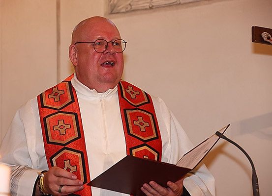Ein Priester steht vor einem Mikrofon und einer Kerze und liest aus einem Buch, wahrscheinlich während eines Gottesdienstes.
