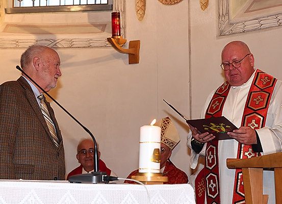 Ein Priester liest ein Buch, während ein anderer Mann neben einer Kerze auf einem Podium steht. Ein Mikrofon und eine Steckdose befinden sich auf dem Tisch.