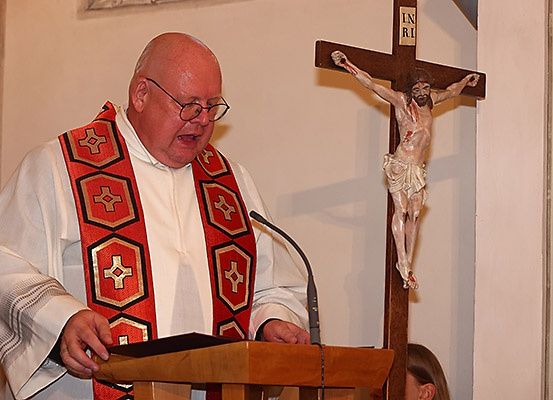 Ein Priester steht an einer Kanzel und trägt ein traditionelles Gewand mit einem Kreuzsymbol. Er spricht in ein Mikrofon. Hinter ihm steht ein Kreuz mit einem Kruzifix.