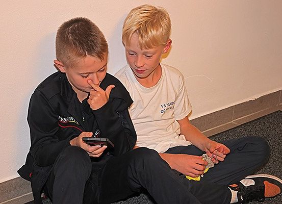 Zwei junge Jungen sitzen auf dem Boden, einer hält ein Handy und der andere ein kleines Objekt. Sie schauen beide auf das Objekt.