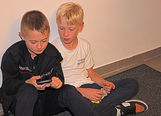 Zwei junge Jungen sitzen auf dem Boden, einer schaut auf ein Handy, der andere hält etwas. Sie tragen Freizeitkleidung und sind von einer weißen Wand umgeben.