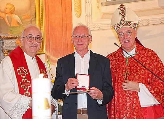 Drei Männer stehen in einer Kirche. Der Mann in der Mitte hält eine rote Schachtel, während die anderen lächeln. Einer trägt einen Bischofshut.