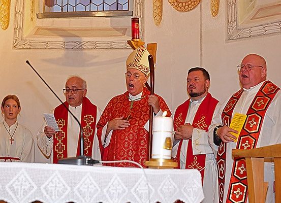 Ein Priester steht während eines Gottesdienstes am Altar, begleitet von zwei anderen Priestern, einer Frau und einem Mikrofon.