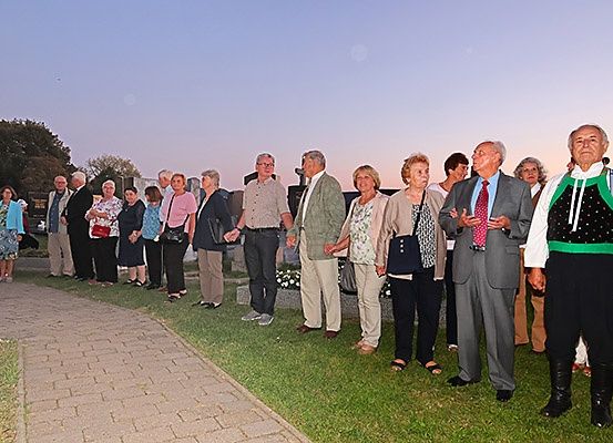 Eine Gruppe von Menschen in formeller Kleidung steht auf einem Rasenfeld, möglicherweise auf einem Friedhof. Sie scheinen in ein Gespräch verwickelt zu sein.