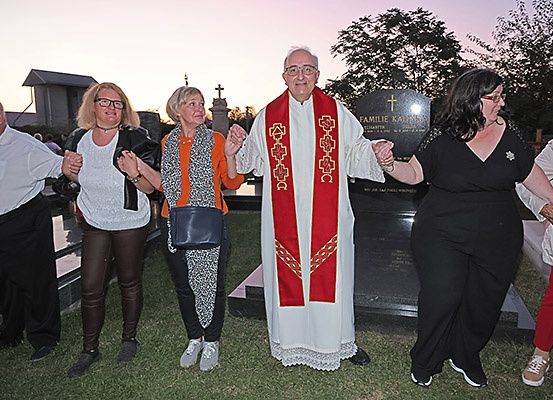 Ein Priester und mehrere Erwachsene stehen um ein Grabmal, möglicherweise bei einer Beerdigungsfeier.