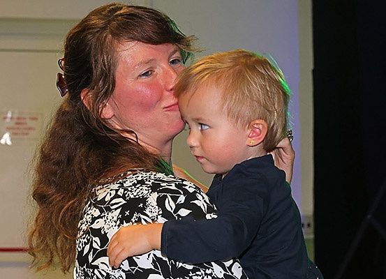 Eine Frau mit langen braunen Haaren küsst einen Jungen mit blonden Haaren auf die Wange, der Junge schaut in die Ferne.