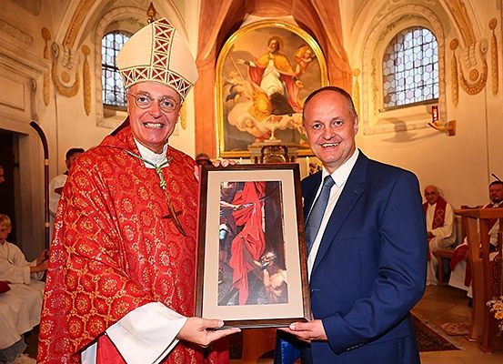 Zwei Männer, einer in geistlicher Kleidung und der andere in einem Anzug, präsentieren ein gerahmtes Kunstwerk. Sie stehen in einer Kirche mit Bogenfenstern und einem Gemälde hinter ihnen.