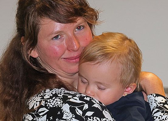 Eine Frau mit langen braunen Haaren hält ein schlafendes Kind mit blonden Haaren. Die Frau trägt ein schwarzes und weißes Blumenhemd.