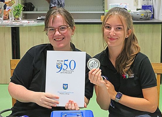Zwei Frauen, eine hält ein Zertifikat und die andere eine Medaille, beide lächelnd, sitzend. Dahinter ein Tresen mit Gegenständen und ein Regal mit verschiedenen Artikeln.