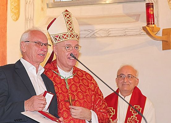 Drei ältere Männer stehen drinnen. Ein Mann trägt eine Mitra und hält ein Mikrofon, ein anderer hält eine rote Schachtel und Papier, und der dritte trägt einen roten Talar.