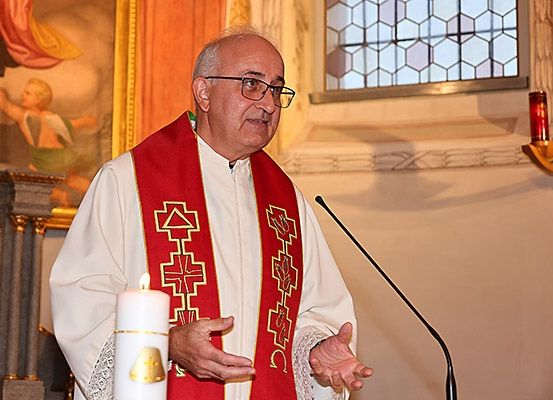 Ein Priester steht in einer Kirche an einer Kanzel, mit einer Kerze und einem Mikrofon. Hinter ihm befindet sich ein religiöses Gemälde, ein Buntglasfenster und ein Kerzenhalter.