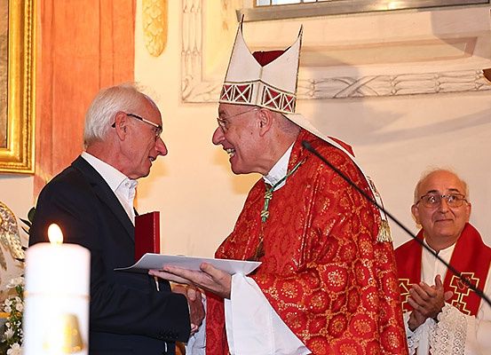 Eine religiöse Zeremonie mit einem Bischof, der einem Mann im Anzug ein Dokument präsentiert, der vor einer Kerze steht und von anderen Priestern flankiert wird.