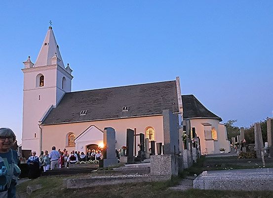 Eine ältere Frau steht in der Nähe einer Kirche mit einem Turm, während sich eine Gruppe für einen Abendgottesdienst versammelt. Grabsteine und Blumen schmücken den Friedhof unter einem blauen Himmel.