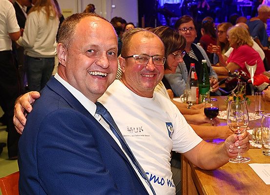Zwei Männer sitzen an einem Tisch in einem Restaurant, lächeln und halten Wein glasses. Hinter ihnen sitzt eine Gruppe von Menschen an einem Tisch. Der Hintergrund ist unscharf, mit stehenden und sitzenden Personen.