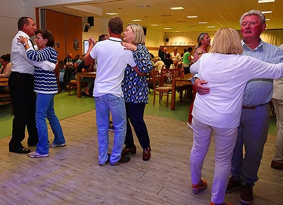 Eine Gruppe von Menschen tanzt in einem Saal mit Holzboden. Einige sind zu zweit, während andere sitzen. Der Raum ist mit Tischen und Stühlen gefüllt.